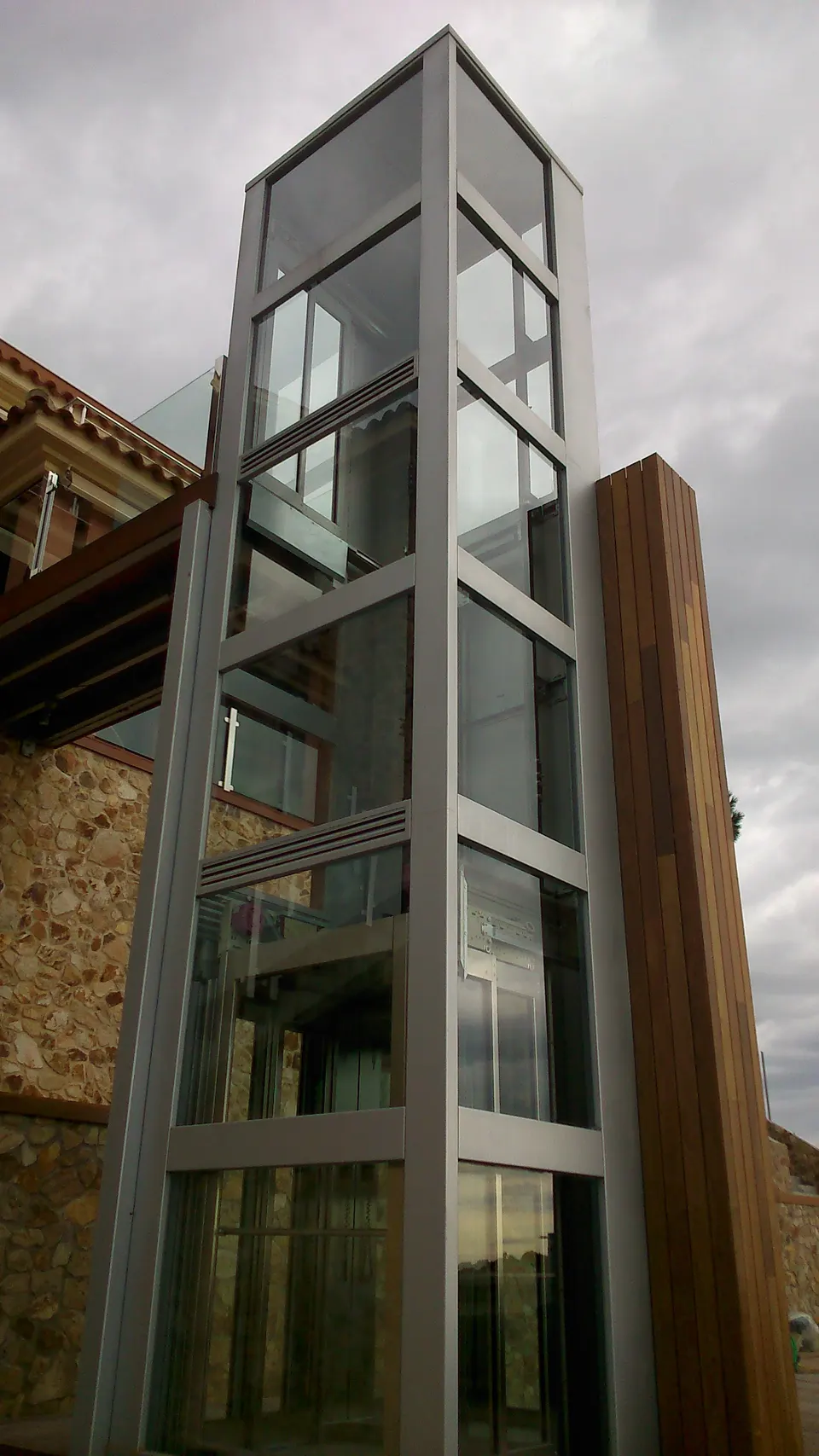 Full-height glass and steel OnLevel EV8 lift structure installed on the exterior of a stone and timber home